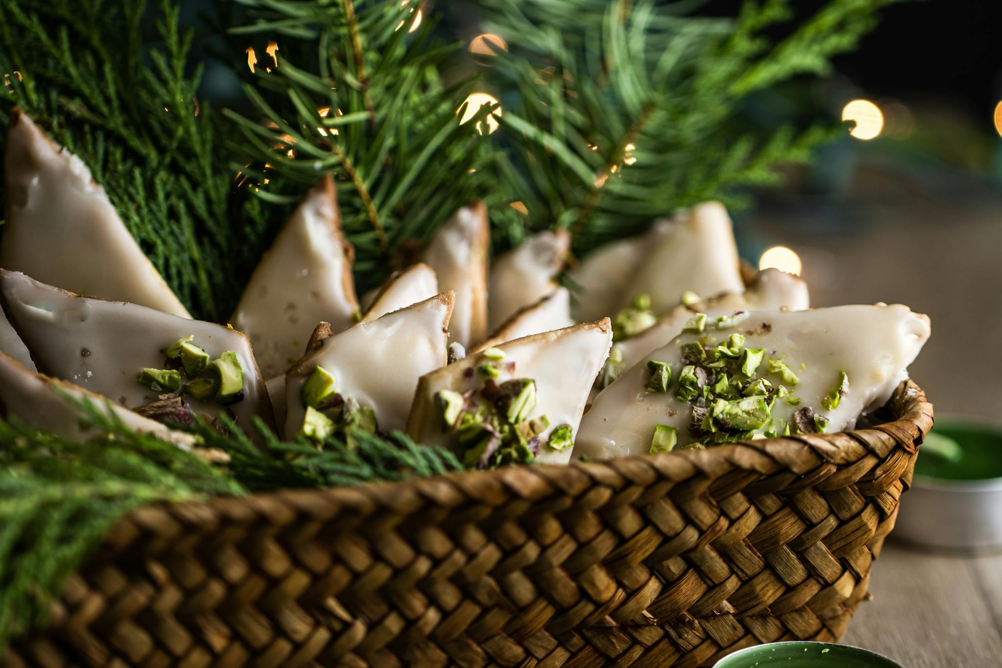 A woven basket filled with glazed triangular cookies topped with pistachios, surrounded by evergreen sprigs.